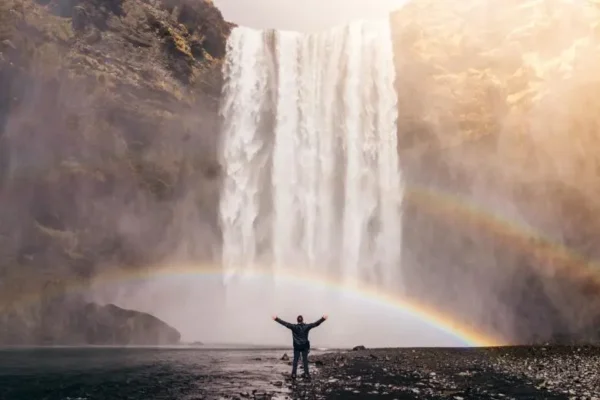 A man stands before a majestic waterfall, a vibrant rainbow arching above, embodying the beauty of nature responsibly. | Don’t Go Chasing Waterfalls: How to Enjoy Nature Responsibly - The Explore Horizon